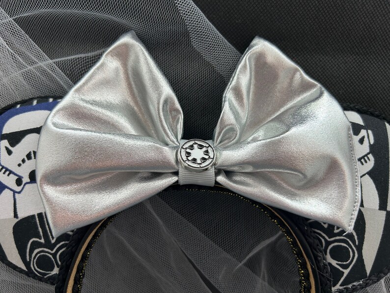May include: Silver bow with a white and silver Imperial symbol button, on a black headband with a white and black Stormtrooper design.