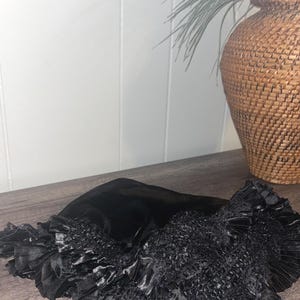 以下が含まれることがあります： 黒色のフリルデザインの布製アクセサリー。光沢のあるサテンのような質感で、ギャザーの入ったテクスチャード加工の中央部があります。縁は幅広でプリーツ加工されています。木製の表面に表示され、背景には織りのバスケットと植物があります。