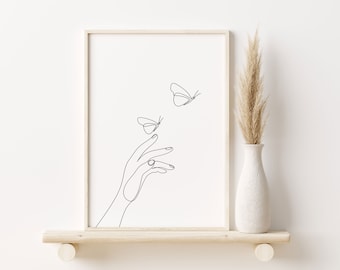 May include: A black and white line art print of a hand reaching out to two butterflies. The print is framed in a light wood frame and is displayed on a shelf with a vase of dried pampas grass.