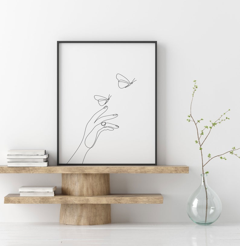 May include: Black and white line art print of a hand reaching out to two butterflies. The print is framed in a black frame and is displayed on a wooden shelf with a vase of greenery.