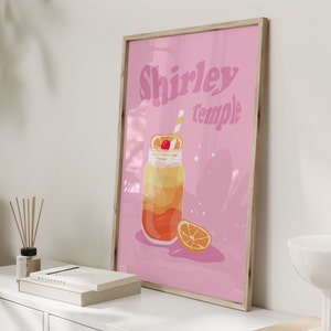 May include: A pink framed poster with the text "Shirley Temple" in a retro font. The poster features a cartoon illustration of a glass jar filled with a layered orange and yellow drink with a cherry on top and a straw. A slice of orange is next to the jar.