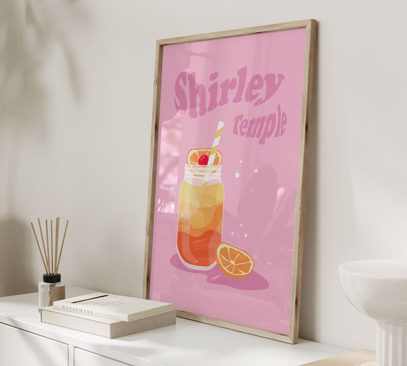 May include: A pink framed poster with the text "Shirley Temple" in a retro font. The poster features a cartoon illustration of a glass jar filled with a layered orange and yellow drink with a cherry on top and a straw. A slice of orange is next to the jar.