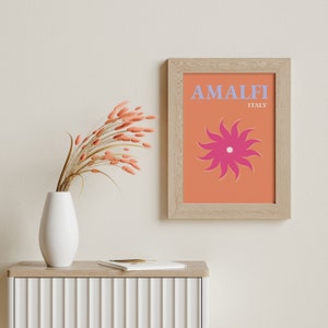 K&ouml;nnte beinhalten: Ein gerahmter Druck mit einem pfirsichfarbenen Hintergrund und einer stilisierten rosa Sonne mit wei&szlig;em Zentrum. Der Text "AMALFI ITALIEN" ist in helllila Buchstaben gedruckt.