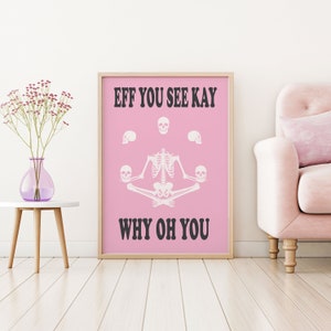 May include: A framed pink poster with the text "EFF YOU SEE KAY WHY OH YOU". The poster features a white skeleton in a yoga pose, surrounded by white skulls. A vase of flowers sits on a white table to the left.