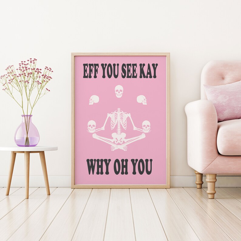 May include: A framed pink poster with the text "EFF YOU SEE KAY WHY OH YOU". The poster features a white skeleton in a yoga pose, surrounded by white skulls. A vase of flowers sits on a white table to the left.