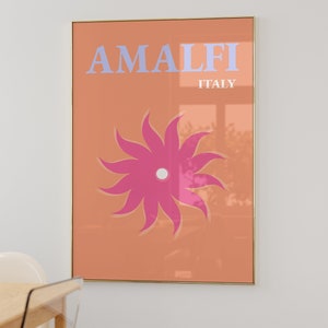 K&ouml;nnte beinhalten: Ein pfirsichfarbener Poster mit goldenem Rahmen zeigt den Text "AMALFI ITALIEN" in hellblauen Buchstaben. Eine stilisierte rosa Blume mit wei&szlig;em Zentrum befindet sich in der Mitte des Posters.