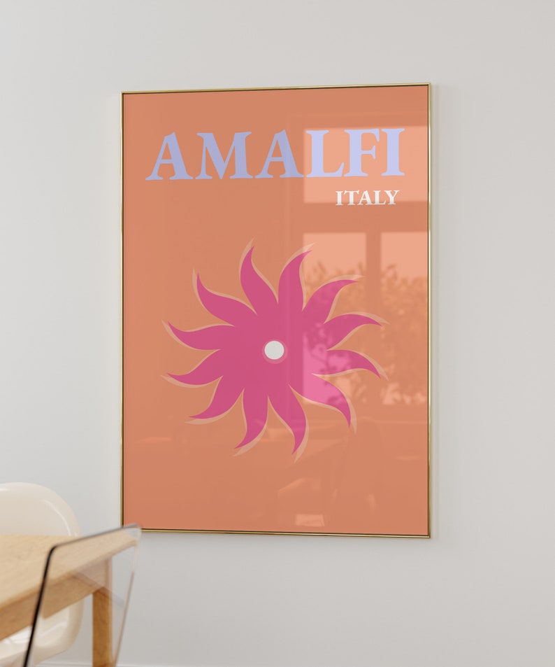 K&ouml;nnte beinhalten: Ein pfirsichfarbener Poster mit goldenem Rahmen zeigt den Text "AMALFI ITALIEN" in hellblauen Buchstaben. Eine stilisierte rosa Blume mit wei&szlig;em Zentrum befindet sich in der Mitte des Posters.
