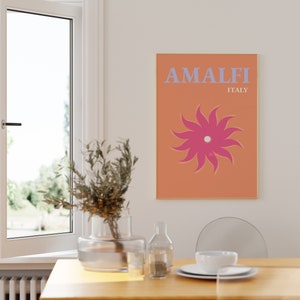 K&ouml;nnte beinhalten: Ein Poster mit einem pfirsichfarbenen Hintergrund und einer rosa Sonne mit einem wei&szlig;en Zentrum. Der Text "AMALFI ITALIEN" ist in blauen Buchstaben.