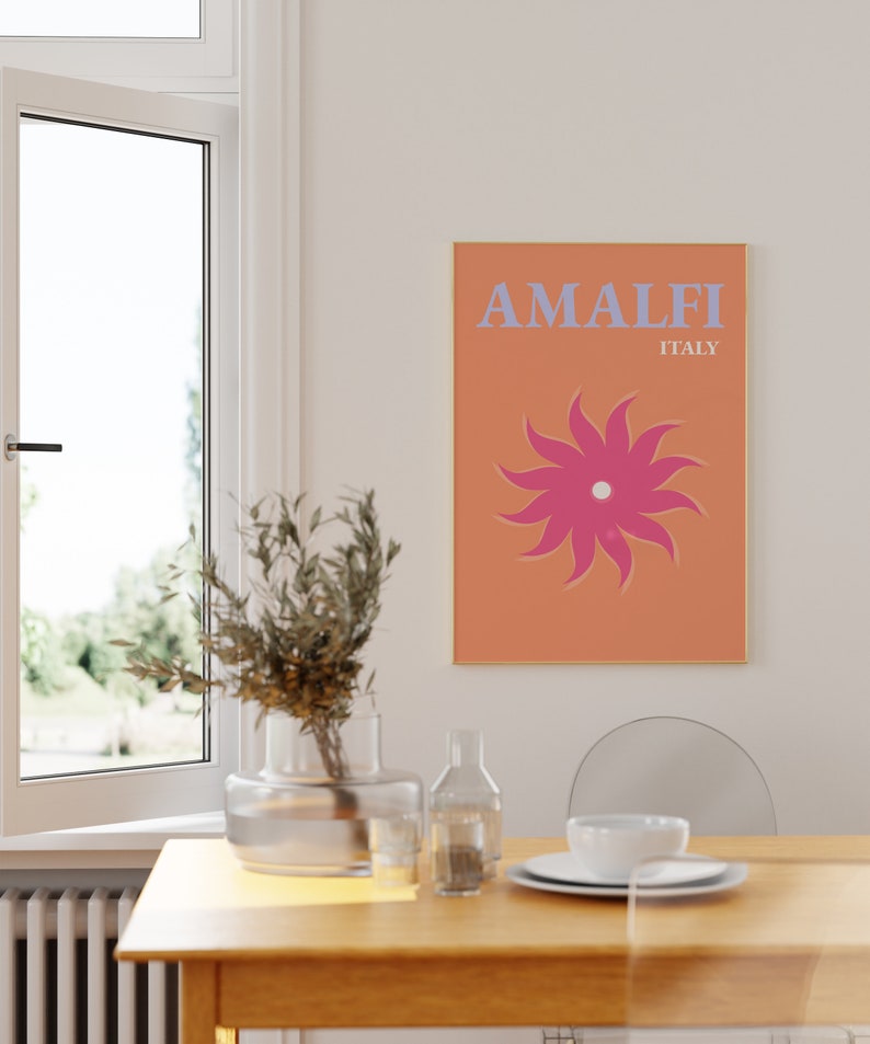 K&ouml;nnte beinhalten: Ein Poster mit einem pfirsichfarbenen Hintergrund und einer rosa Sonne mit einem wei&szlig;en Zentrum. Der Text "AMALFI ITALIEN" ist in blauen Buchstaben.