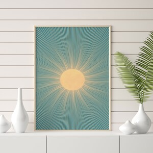 May include: A framed print of a stylized sun with yellow rays on a teal background. The print is hanging on a white wall with a white shelf below it.