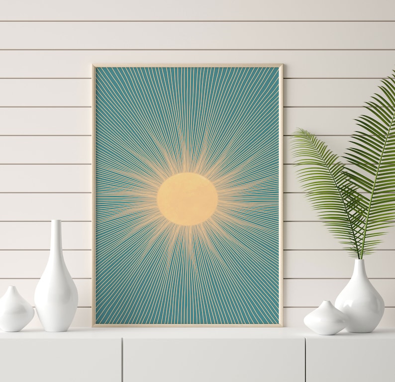May include: A framed print of a stylized sun with yellow rays on a teal background. The print is hanging on a white wall with a white shelf below it.