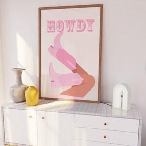 Pu&ograve; includere: Un poster incorniciato con la parola "HOWDY" in lettere rosa e un paio di stivali da cowboy rosa con una gamba marrone.