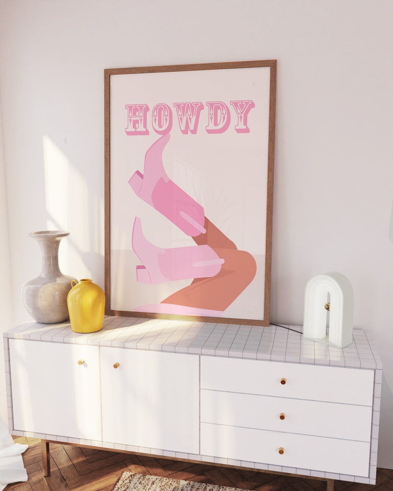 Pu&ograve; includere: Un poster incorniciato con la parola "HOWDY" in lettere rosa e un paio di stivali da cowboy rosa con una gamba marrone.