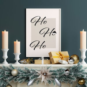 May include: A white framed print with the text "Ho Ho Ho" on a beige background. The print is on a white mantel with a green garland, gold and silver ornaments, and two white candle holders with lit candles.