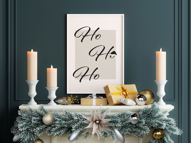 May include: A white framed print with the text "Ho Ho Ho" on a beige background. The print is on a white mantel with a green garland, gold and silver ornaments, and two white candle holders with lit candles.