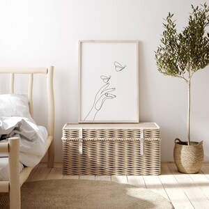 May include: A black and white line art print of a hand reaching out to two butterflies. The print is framed in a light wood frame and is hanging on a white wall above a wicker storage trunk.