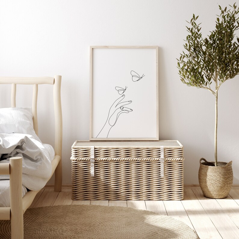 May include: A black and white line art print of a hand reaching out to two butterflies. The print is framed in a light wood frame and is hanging on a white wall above a wicker storage trunk.