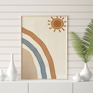 Op de afbeelding: Een ingelijst print met een minimalistisch ontwerp met een gestileerde regenboog in tinten bruin, blauw en beige met een zon in de rechterbovenhoek.