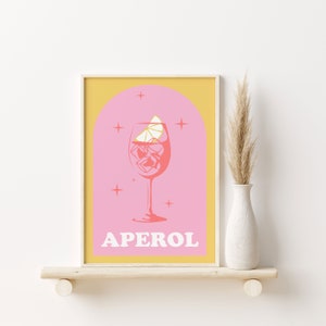 Puede incluir: Una impresi&oacute;n de arte imprimible rosa y amarilla con un vaso de Aperol Spritz con una rodaja de lim&oacute;n y cubitos de hielo. El texto "APEROL" est&aacute; impreso en blanco en la parte inferior de la impresi&oacute;n.