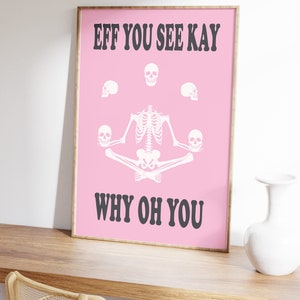 May include: A framed pink poster with the text "EFF YOU SEE KAY WHY OH YOU" in black. The poster features a white skeleton in a yoga pose, surrounded by white skulls. The poster is displayed on a wooden surface.