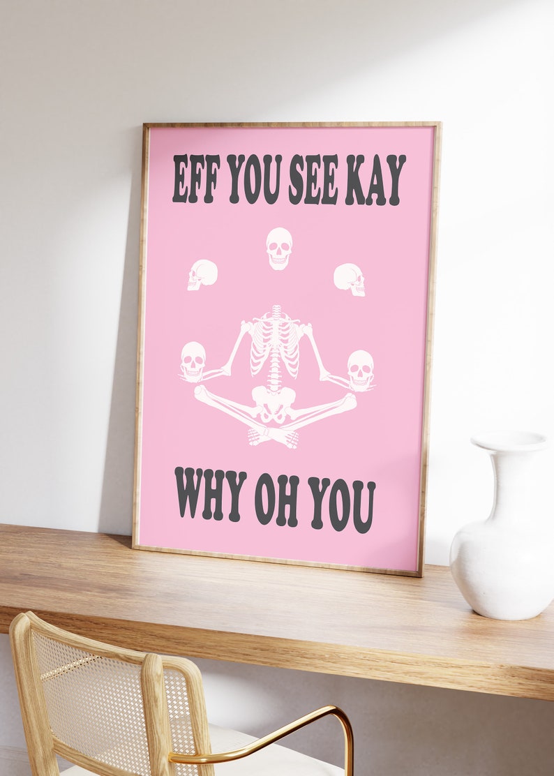 May include: A framed pink poster with the text "EFF YOU SEE KAY WHY OH YOU" in black. The poster features a white skeleton in a yoga pose, surrounded by white skulls. The poster is displayed on a wooden surface.