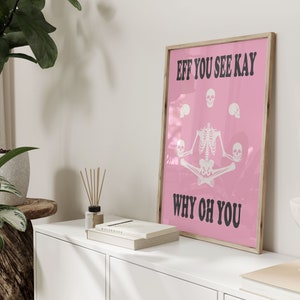 May include: A framed pink poster with the text "EFF YOU SEE KAY WHY OH YOU" in black. The poster features a white skeleton in a seated yoga pose, surrounded by white skulls. The frame is a light wood color.