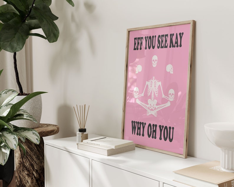 May include: A framed pink poster with the text "EFF YOU SEE KAY WHY OH YOU" in black. The poster features a white skeleton in a seated yoga pose, surrounded by white skulls. The frame is a light wood color.