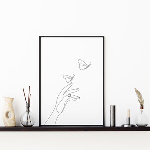 May include: A black and white line drawing of a hand reaching out to two butterflies. The butterflies are flying away from the hand. The drawing is framed in a black frame and is hanging on a white wall.