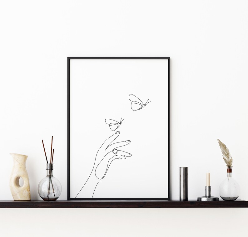 May include: A black and white line drawing of a hand reaching out to two butterflies. The butterflies are flying away from the hand. The drawing is framed in a black frame and is hanging on a white wall.