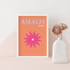 K&ouml;nnte beinhalten: Ein wei&szlig;er gerahmter Druck mit orangefarbenem Hintergrund und einer stilisierten rosa Blume. Der Text "AMALFI ITALIEN" ist in Blau auf dem Druck gedruckt.