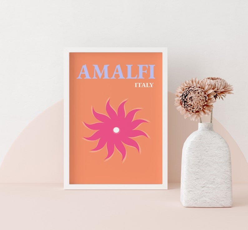 K&ouml;nnte beinhalten: Ein wei&szlig;er gerahmter Druck mit orangefarbenem Hintergrund und einer stilisierten rosa Blume. Der Text "AMALFI ITALIEN" ist in Blau auf dem Druck gedruckt.