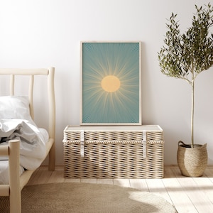 May include: A wicker storage trunk with a light wood frame and a white canvas top. The trunk is sitting in front of a white wall with a framed print of a sun with teal rays. A potted olive tree is on the right side of the image.