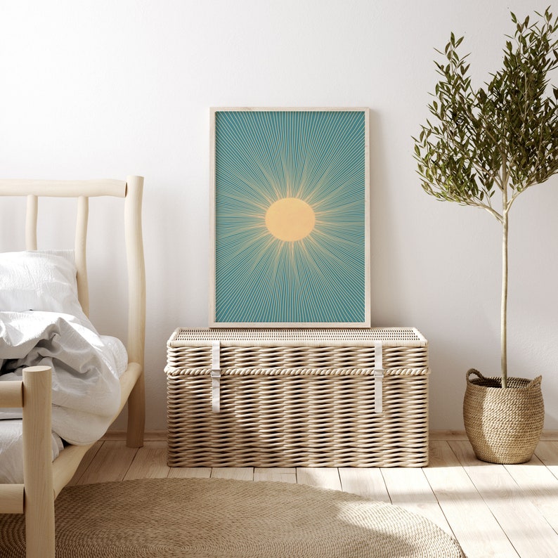 May include: A wicker storage trunk with a light wood frame and a white canvas top. The trunk is sitting in front of a white wall with a framed print of a sun with teal rays. A potted olive tree is on the right side of the image.