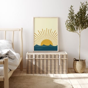 May include: A framed print of a stylized sun rising over blue waves. The sun is yellow and the waves are a deep blue. The background is a light yellow.