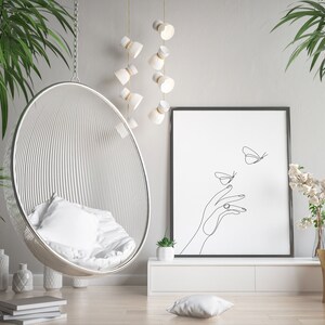 May include: A white hanging chair with a white cushion, a black framed print with a line drawing of a hand and butterflies, a white shelf with a small potted plant, and a wooden stump with a vase of white flowers.