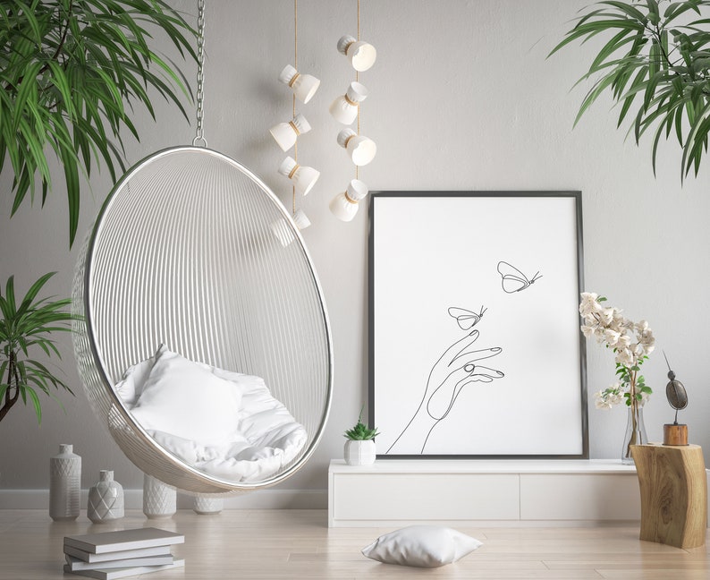 May include: A white hanging chair with a white cushion, a black framed print with a line drawing of a hand and butterflies, a white shelf with a small potted plant, and a wooden stump with a vase of white flowers.