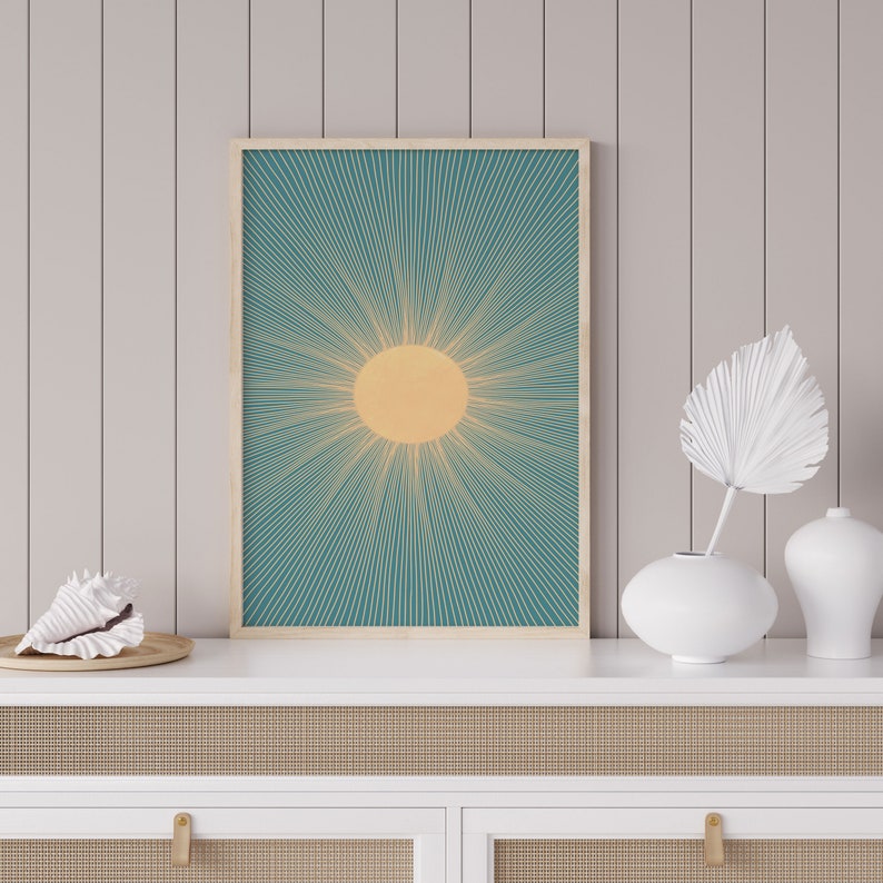 May include: A teal and yellow sunburst print in a wooden frame hangs above a white dresser with two drawers. The dresser has a woven rattan front.