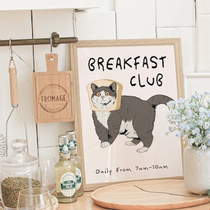 Könnte beinhalten: Ein weißer gerahmter Druck mit dem Text "Breakfast Club" und einer Cartoon-Darstellung einer grauen Katze, die eine Scheibe Brot als Hut trägt. Der Text "Daily From 7am-10am" befindet sich unter dem Bild.