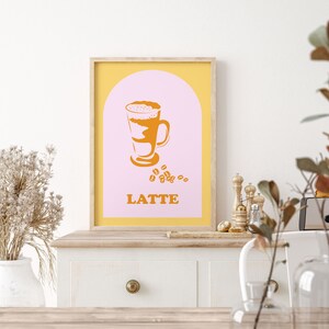 May include: A printable poster with a yellow frame and a pink background. The poster features a stylized illustration of a latte in a glass with a brown and white design. The text "LATTE" is printed in orange below the illustration.