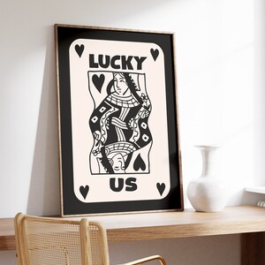 Pu&ograve; includere: Stampa in bianco e nero di una carta da gioco della Regina di Cuori con il testo "LUCKY US" su uno sfondo rosa.