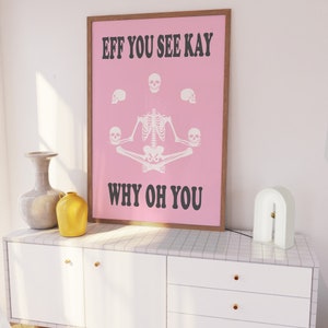 May include: A framed pink poster with the text "EFF YOU SEE KAY WHY OH YOU" and a skeleton illustration. The poster is in a brown frame and sits on a white cabinet with gold knobs. A white lamp and vases are also on the cabinet.