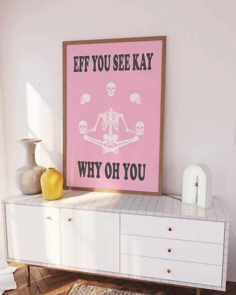 May include: A framed pink poster with the text "EFF YOU SEE KAY WHY OH YOU" and a skeleton illustration. The poster is in a brown frame and sits on a white cabinet with gold knobs. A white lamp and vases are also on the cabinet.