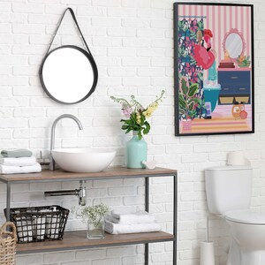 May include: A white bathroom with a white toilet, a white sink with a chrome faucet, a round mirror with a black frame, a wooden shelf with towels, a wicker basket, and a framed print of a woman in a bathroom with a pink flamingo.
