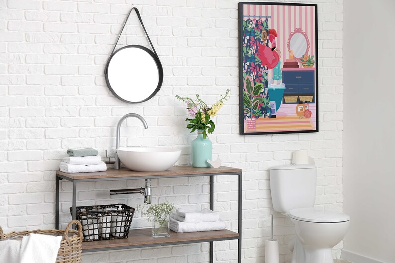May include: A white bathroom with a white toilet, a white sink with a chrome faucet, a round mirror with a black frame, a wooden shelf with towels, a wicker basket, and a framed print of a woman in a bathroom with a pink flamingo.