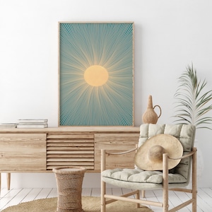 May include: A teal and yellow sunburst print in a wooden frame. The print is hanging on a white wall above a wooden cabinet and a wicker stool. A chair with a straw hat on it is in the foreground.
