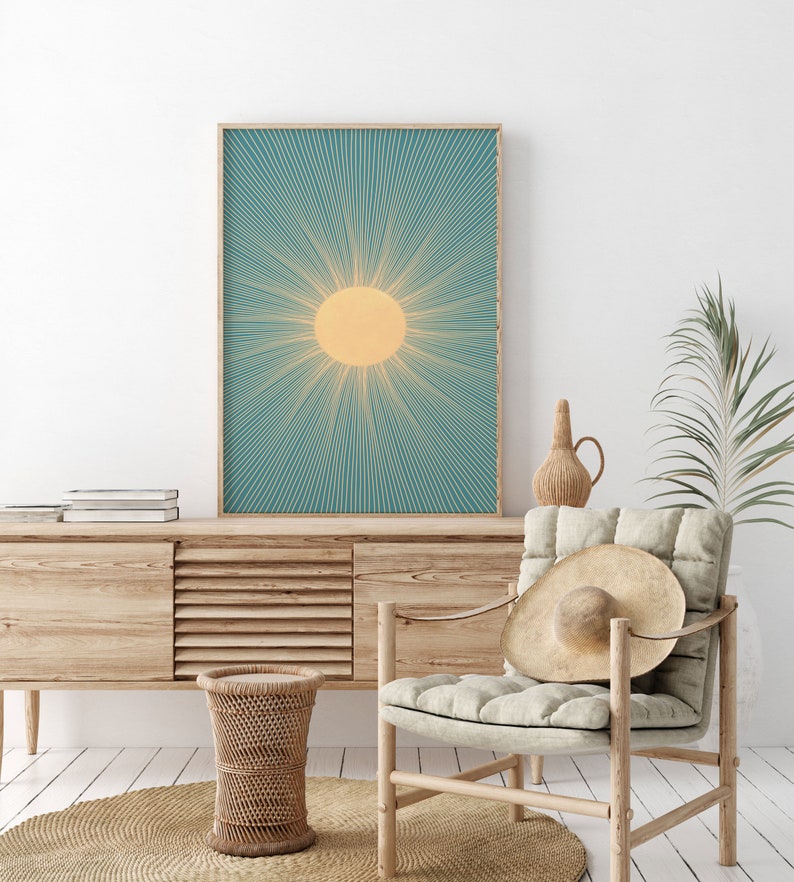 May include: A teal and yellow sunburst print in a wooden frame. The print is hanging on a white wall above a wooden cabinet and a wicker stool. A chair with a straw hat on it is in the foreground.