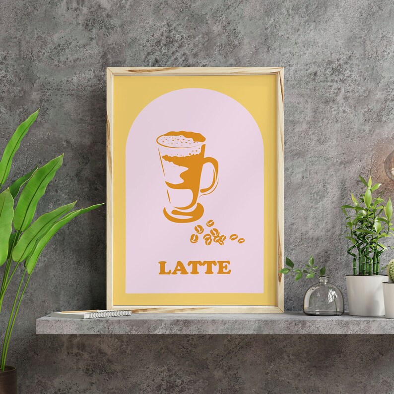May include: A framed print with a yellow background and a pink arch. The arch contains a stylized illustration of a latte in a glass with coffee beans scattered below. The text "LATTE" is printed in orange below the illustration.