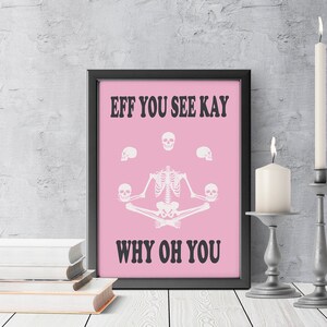 May include: A framed pink print with the text "EFF YOU SEE KAY WHY OH YOU" and a skeleton in a yoga pose. The print is in a black frame, with two lit candles in silver candle holders and a stack of books.