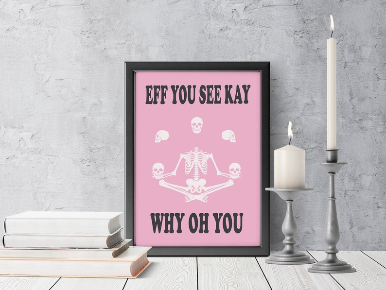 May include: A framed pink print with the text "EFF YOU SEE KAY WHY OH YOU" and a skeleton in a yoga pose. The print is in a black frame, with two lit candles in silver candle holders and a stack of books.
