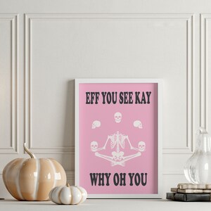May include: A framed pink print with white text and a skeleton illustration. The text reads "EFF YOU SEE KAY WHY OH YOU". The skeleton is in a yoga pose, holding skulls. Decorative pumpkins and candles are in the foreground.